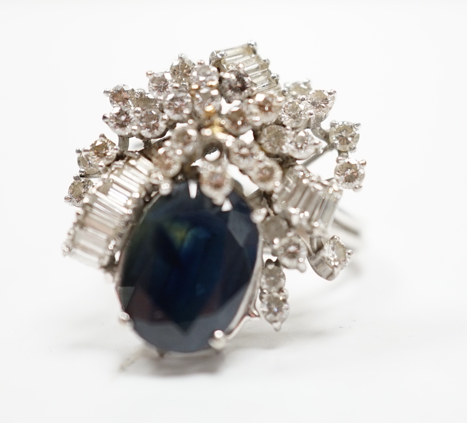 A mid to late 20th century 18k white metal, single stone oval cut sapphire and baguette and round cut diamond cluster set dress ring, size M/N, gross weight 10.3 grams.